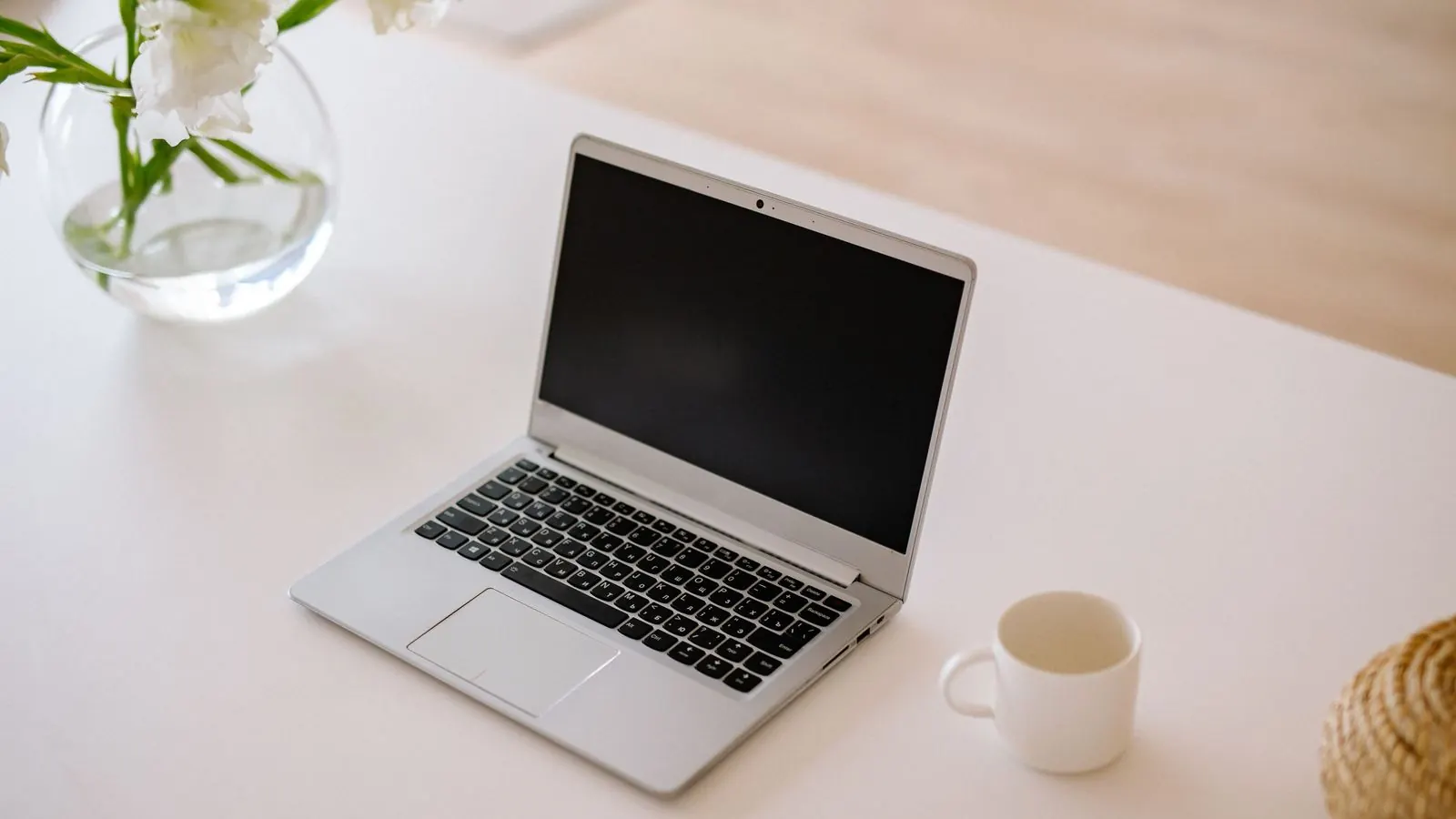 Otwarty laptop i biała filiżanka na jasnym stole, obok szklany wazon z białymi kwiatami
